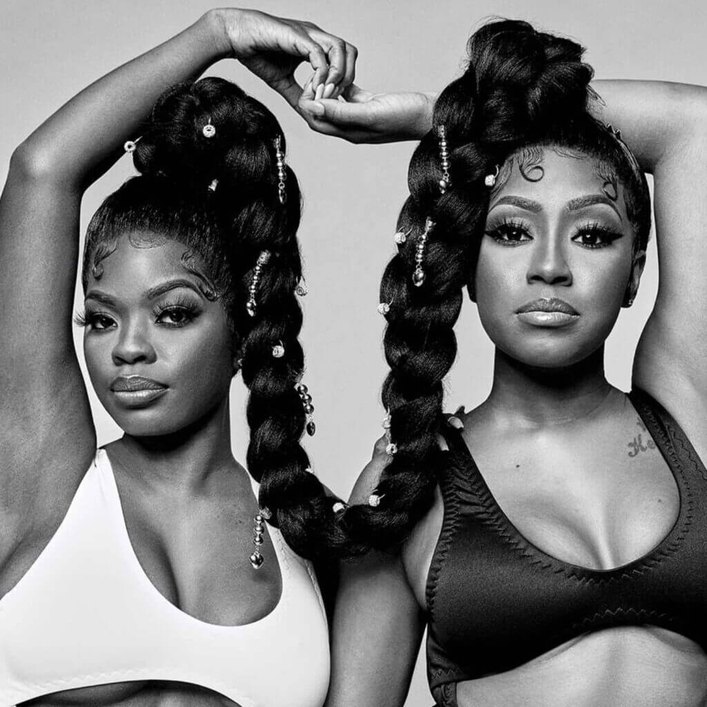 Two women, capturing a No Mercy vibe, showcase long braided hairstyles while posing in sports bras. The woman on the left is in a white bra with her arms raised overhead, and the one on the right wears a black bra, gracefully resting one hand on her shoulder. The photograph is presented in black and white.