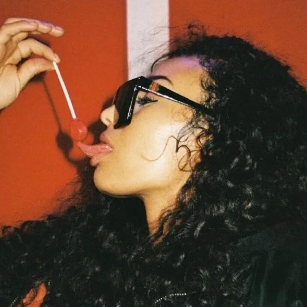 A person sporting curly hair and sunglasses enjoys a Watchu Want lollipop set against a vivid red backdrop.