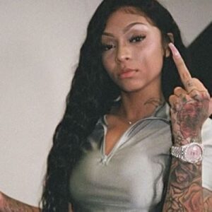 A person with long wavy hair and tattooed arms is showcasing a Bossin silver-tone watch while wearing a light-colored top, posing with a neutral expression and one hand raised.