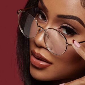 A close-up shot featuring a person in round glasses, adorned with long lashes and manicured nails, showcasing a sense of confidence while embodying the "Clumsy" allure. Their face is set against a solid maroon background.