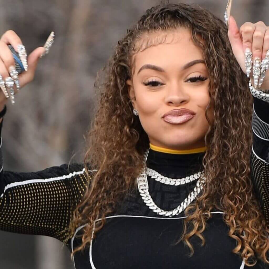 The scene captures someone with curly hair confidently showcasing vibrant energy in an all-black ensemble featuring yellow accents. Their ornate, long nails stand out as they gesture with both hands raised, accentuated by layered silver necklaces. The background is artfully blurred, drawing focus to their dynamic presence and the essence of "Energy.