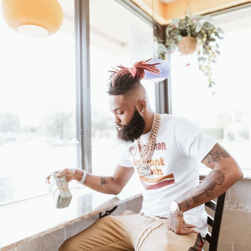 A man with a beard and braided hair sits by a window, wearing a white t-shirt and gold chains, holding a stack of cash. The bright light filters through the window, casting patterns across his face reminiscent of the Paper Trail design, with a hanging plant visible in the background.